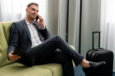 Cheerful Businessman Using Phone Sitting In Hotel Room With Suitcase On Business Trip Stock