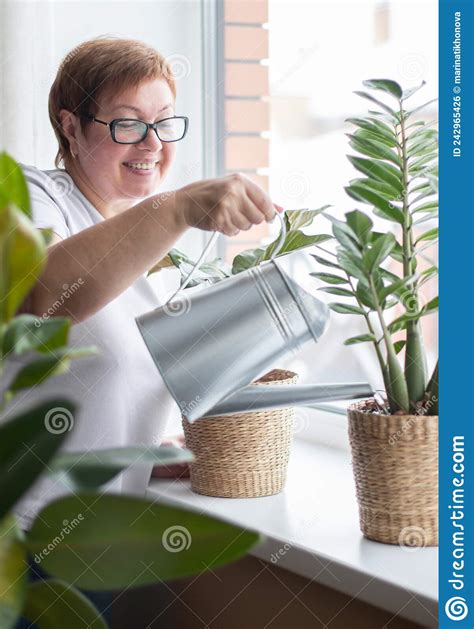 An Elderly Woman Waters Home Plants On The Windowsill From A Watering