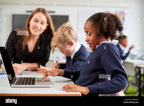 A Female School Teacher Sitting At A Desk Helping A Boy And Girl Using