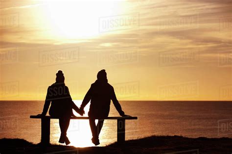 Silhouette Of Couple Sitting On Bench Holding Hands Against Sunset