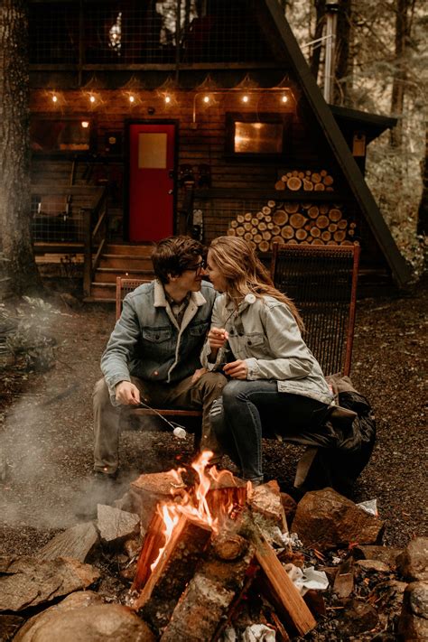 A Man And Woman Sitting Next To A Campfire In Front Of A Log Cabin