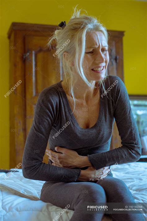 Mature Woman Sitting On Bed Clutching Stomach In Pain Interior Blonde Stock Photo