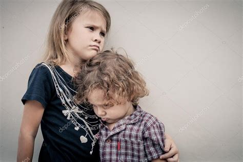 Portrait Of Sad Little Girl And Little Boy Standing Near Wall In