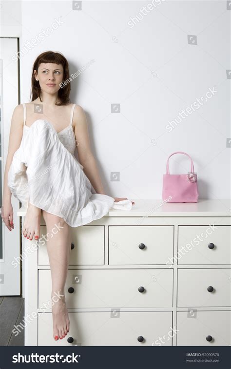 Smiling Flirting Woman Wearing White Dress Sitting On Chest Of Drawers