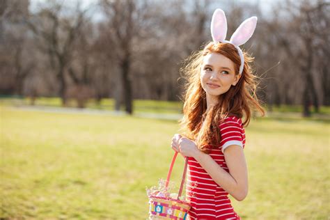 portrait of a smiling cute red head girl wearing bunny ears and holding basket full of easter