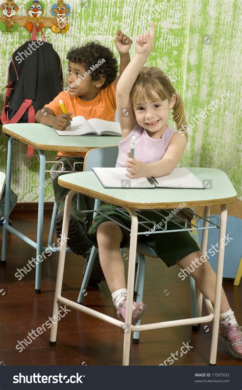 Two Students Sitting At Desks In A Classroom They Have Their Hands