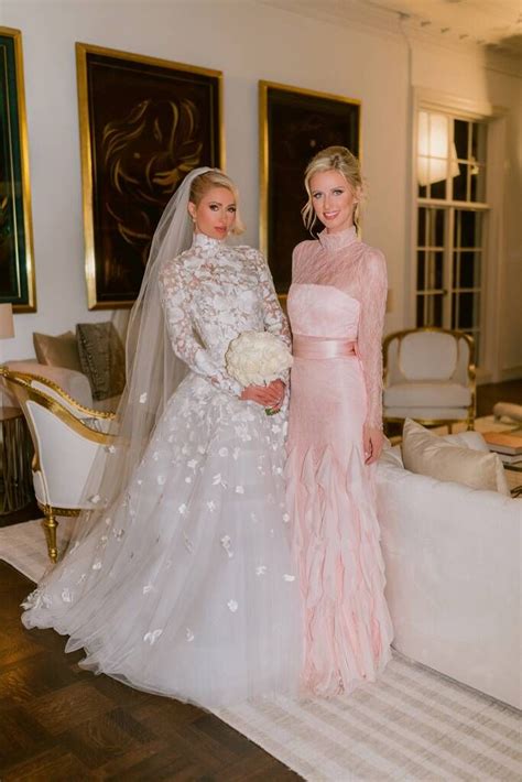 Two Women In Wedding Gowns Standing Next To Each Other Near A Couch With Flowers On It