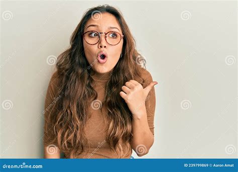 Young Hispanic Girl Wearing Casual Clothes And Glasses Surprised Pointing With Hand Finger To