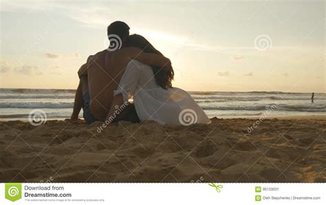 A Woman And A Man Sits Together In The Sand On The Sea Shore Admiring The Ocean And Landscapes