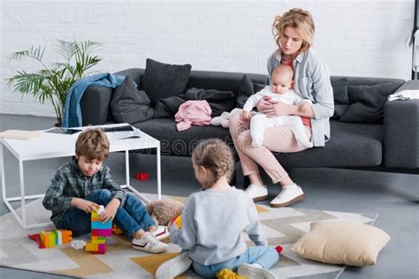 Tired Mother With Infant Child Sitting On Sofa And Looking At Siblings