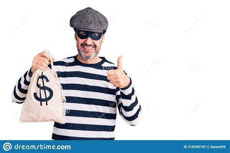 middle age handsome man wearing burglar mask holding money bag smiling happy and positive thumb