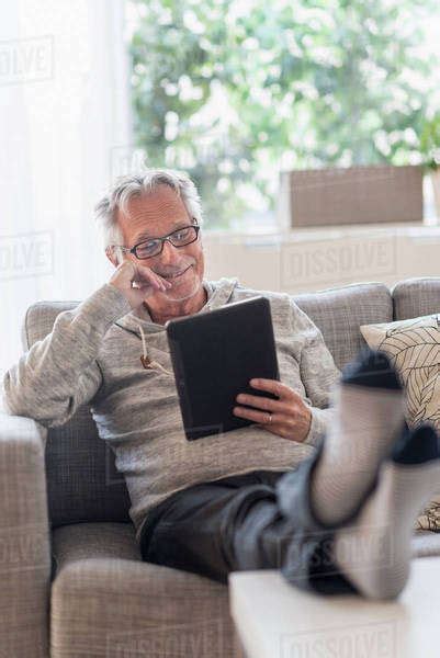 Senior Man Sitting On Couch In Living Room Using Tablet Pc And Smiling