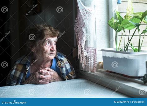 A Lonely Old Woman Is Sitting By The Window In Her House Stock Photo
