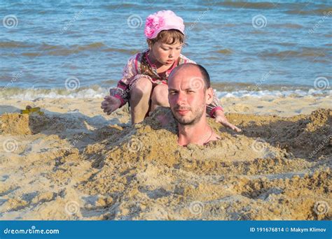 The Head Of A Man Buried In The Sand Is Next To A Little Girl On The