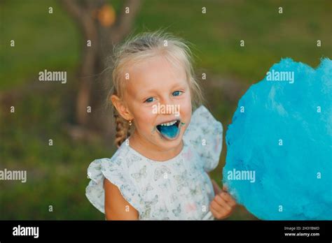Little Blond Girl Eating Cotton Candy And Shows Blue Tongue In The Park