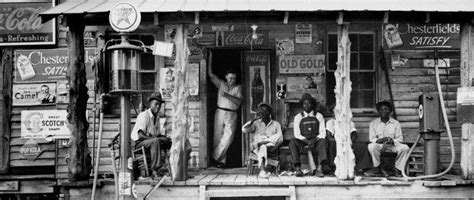 An Old Black And White Photo Of People Sitting On The Porch Of A Storefront