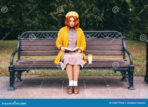 A Beautiful Red Haired Girl With A Curly Hairstyle Is Sitting On A Park