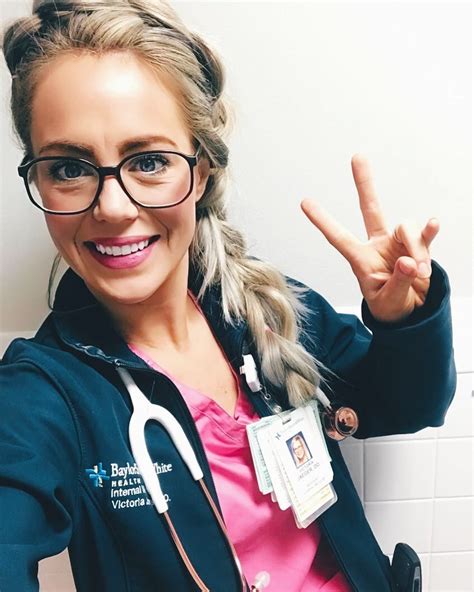 A Woman With Glasses And A Nurses Coat Is Making The Peace Sign While Standing In Front Of A