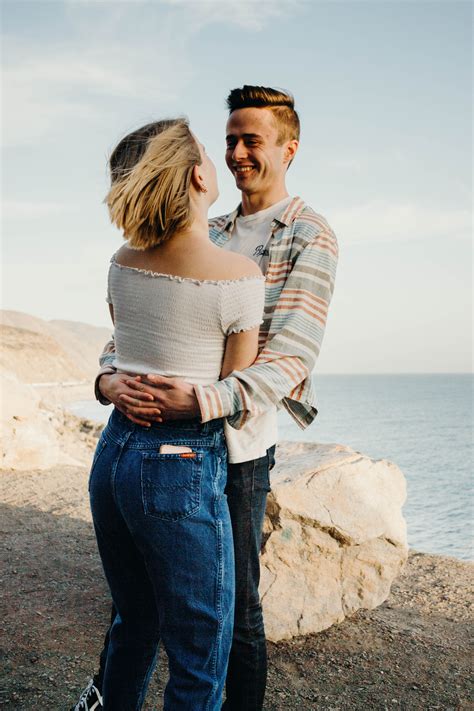 A Man And Woman Standing Next To Each Other Near The Ocean With Their Arms Around Each Other