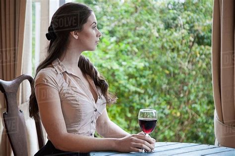 Young Woman Sitting At Table In Front Of Window Holding Glass Of Wine