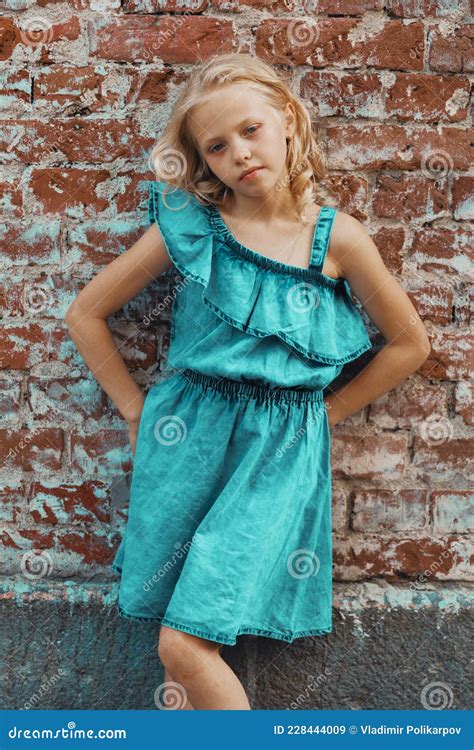 A Young Blonde Girl In A Blue Dress Poses Against A Brick Wall Stock