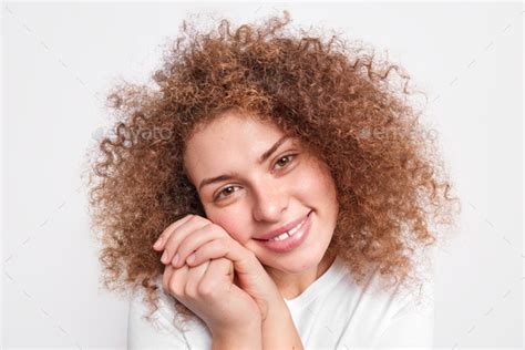 portrait of good looking curly haired teenage girl tilts head keeps hands near face smiles