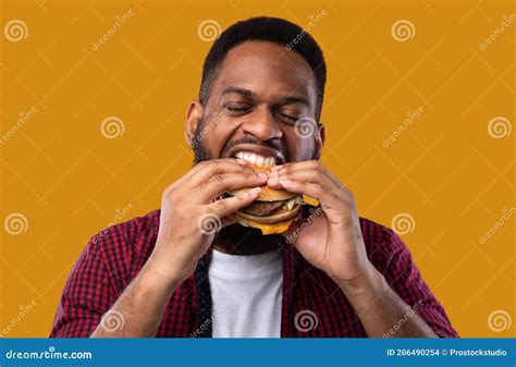 Hungry African American Guy Eating Burger Standing On Yellow Background