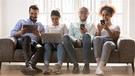 Smiling Diverse Young People Sit On Couch Using Gadgets Stock Photo