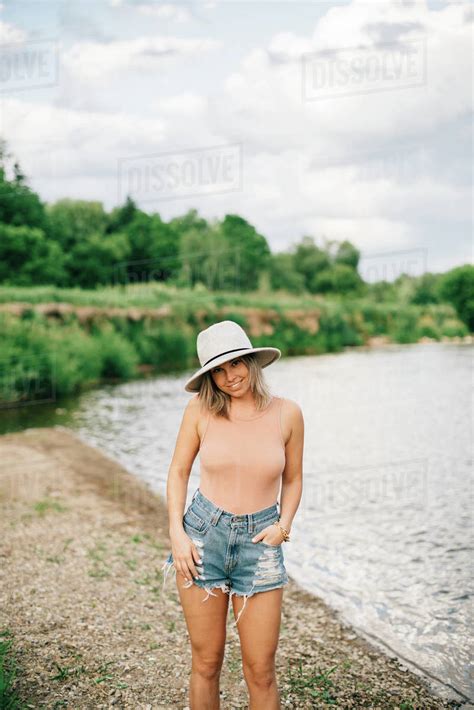 Babe Blonde Woman Wearing Shorts And Hat Standing On A Riverbank Smiling At Camera Stock