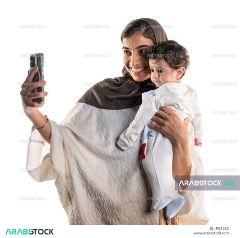 بورتريه لأم عربية خليجية سعودية تحمل طفلها الرضيع بسعادة وحنان، تقوم بإلتقاط صورة سلفي تذكارية