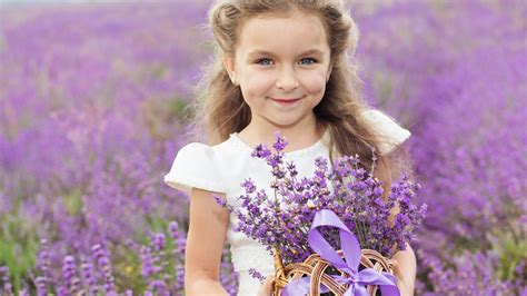 Smiling Cute Little Girl With Purple Flowers Basket Is Standing In Blur
