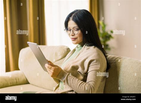mature asian woman wearing glasses is deeply focused on browsing the internet on her tablet