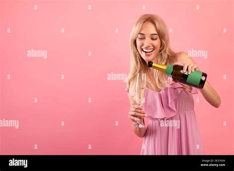 Cheerful Blonde Lady Pouring Champagne Into Glass Over Pink Studio