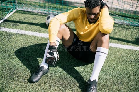 African American Goalkeeper Sitting At Goalpost On Pitch Stock Photo