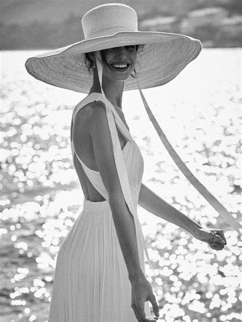A Woman In A White Dress And Wide Brimmed Hat Smiles At The Camera