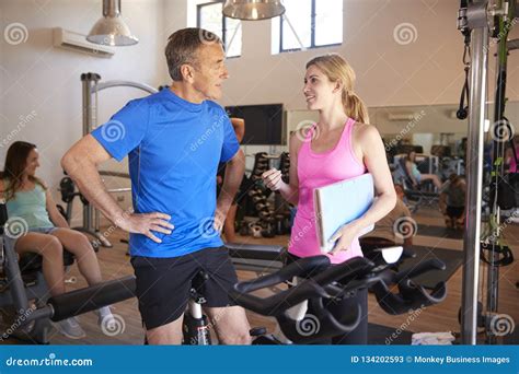 Senior Man Exercising On Cycling Machine Being Encouraged By Female