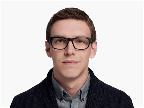 a man wearing glasses and a sweater looks at the camera while standing in front of a white