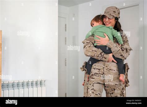Female Soldier In Camouflage Uniform And Cap Carrying Anonymous Barefoot Son In Light Room At