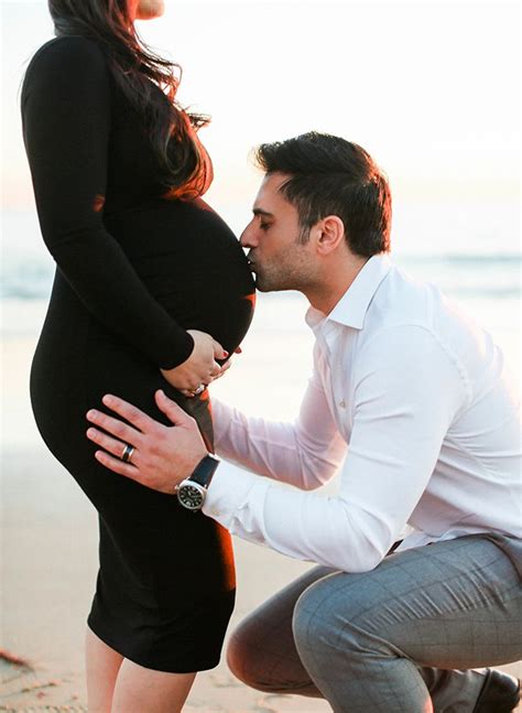 A Man Kneeling Down To Kiss A Pregnant Womans Belly On The Beach At Sunset