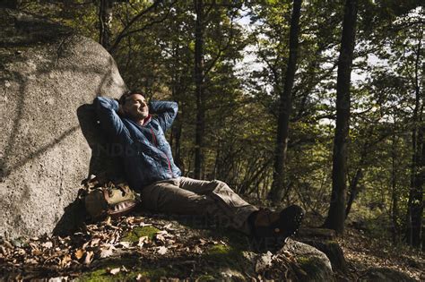 Mature Man With Hands Behind Head Relaxing By Rock In Forest Stock Photo