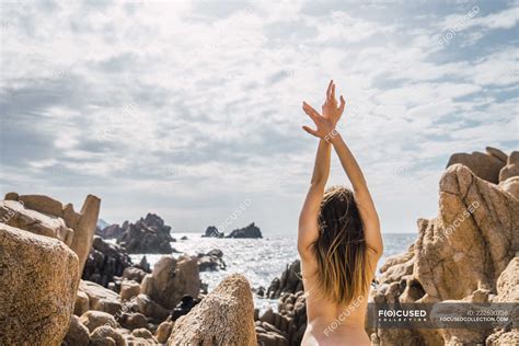 Back View Of Naked Woman With Hands Up Standing And Stretching At Coastal Rocks At The Ocean