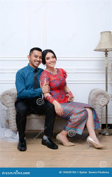 portrait of beautiful happy indonesian couple wearing traditional costume sitting on couch stock