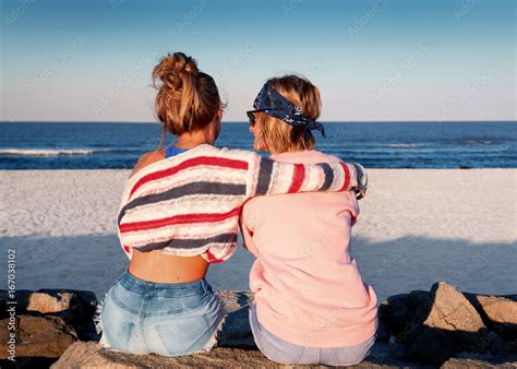 Two Young Girls Best Friends Sitting Together On The Beach At Sunset