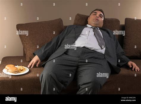 Obese Man In Formal Clothes Sleeping On Sofa With Plate Of Burgers And