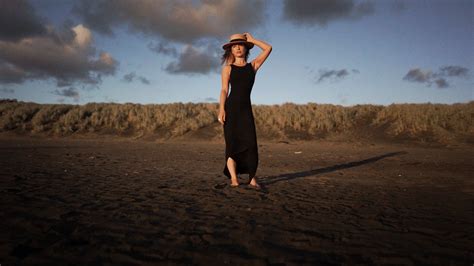 A Woman In A Black Dress And Hat Standing On The Beach With Her Hands Behind Her Head