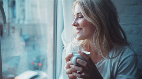 Portrait Of Charming Young Blonde Woman Sitting On Windowsill Drink