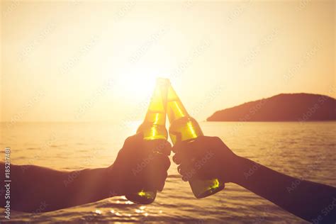 Couple Hands Holding Beer Bottles And Clanging Celebrating On Holiday At The Beach In Sunset
