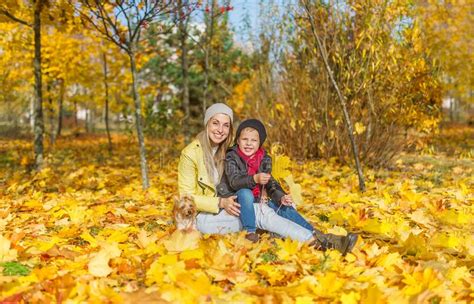 Portrait Of Mother And Son Sitting In The Autumn Park Among The Falling