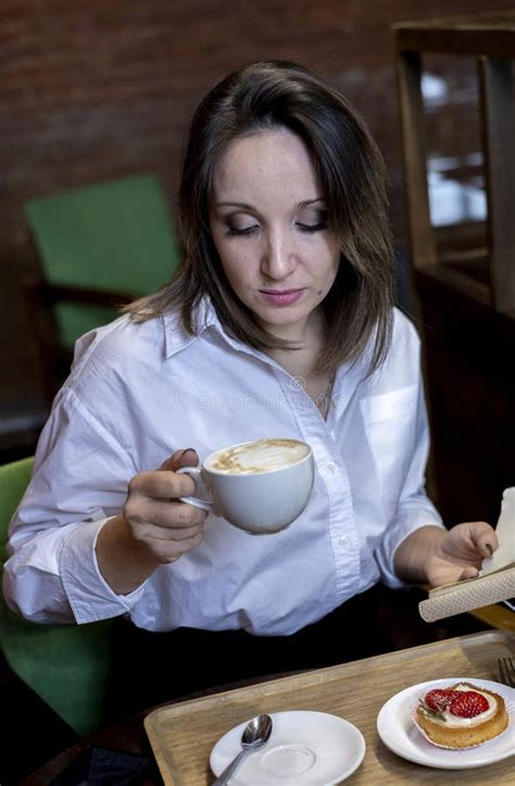 Young Woman In White Shirt Sits In Cafe And Holds Cup Of Coffee In