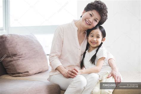 Chinese Grandmother And Granddaughter Hugging On Sofa And Looking In